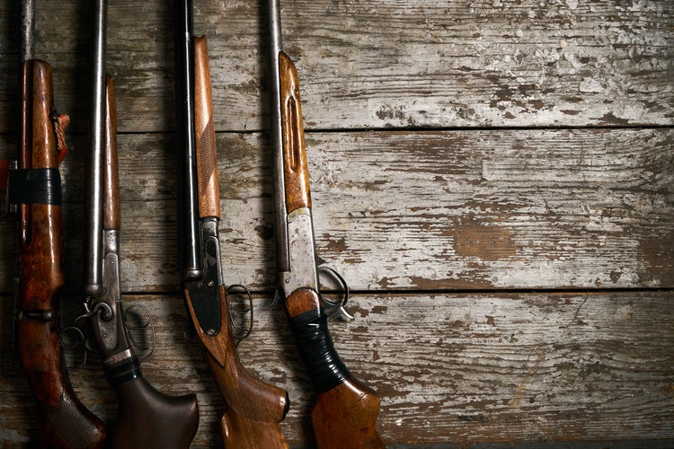 Four vintage rifles with wooden stocks are lined up vertically against a worn, rustic wooden background.